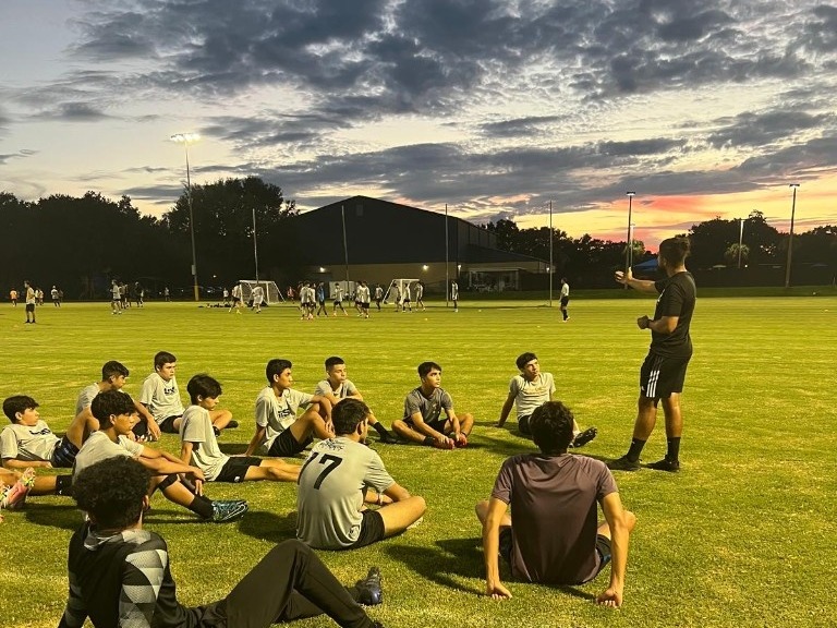 Mauricio talking with a youth soccer team on the field at sunset
